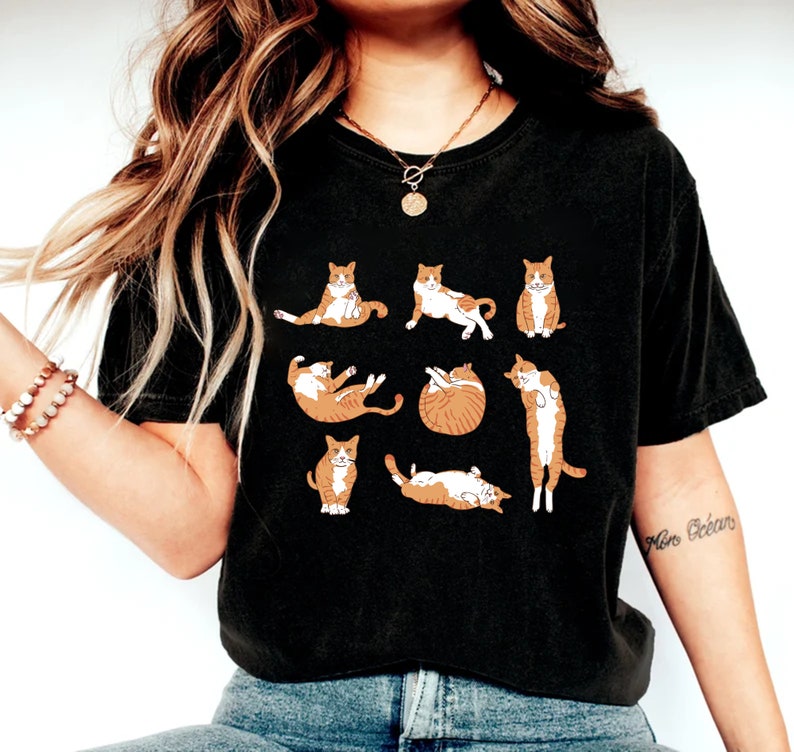 May include: Black t-shirt featuring a pattern of orange and white cats in various poses. The cats are depicted sitting, stretching, and curled up. The shirt is a comfortable, casual style.