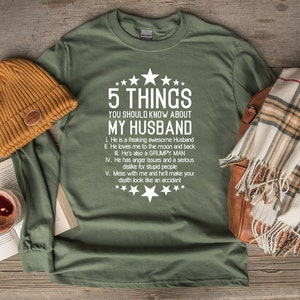 May include: Olive green long-sleeve shirt with the text "5 THINGS YOU SHOULD KNOW ABOUT MY HUSBAND" in white. The shirt is paired with a mustard-colored beanie, a plaid scarf, and a leather bag.