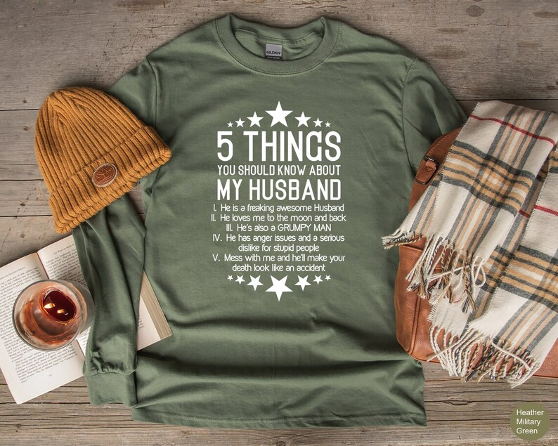 May include: Olive green long-sleeve shirt with the text "5 THINGS YOU SHOULD KNOW ABOUT MY HUSBAND" in white. The shirt is paired with a mustard-colored beanie, a plaid scarf, and a leather bag.