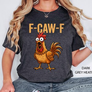 May include: Dark grey heather t-shirt featuring the text "F-CAW-F" in gold above a cartoon rooster with an open beak. The rooster is brown, orange, and red.