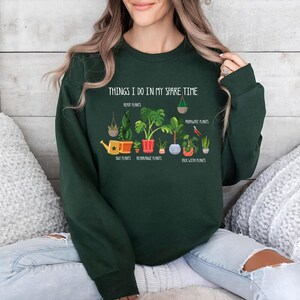 May include: Forest green sweatshirt with the text "THINGS I DO IN MY SPARE TIME" and illustrations of plants. The graphic includes text such as "Repot Plants", "Buy Plants", "Repot Plants", "Propagate Plants", and "Talk With Plants".