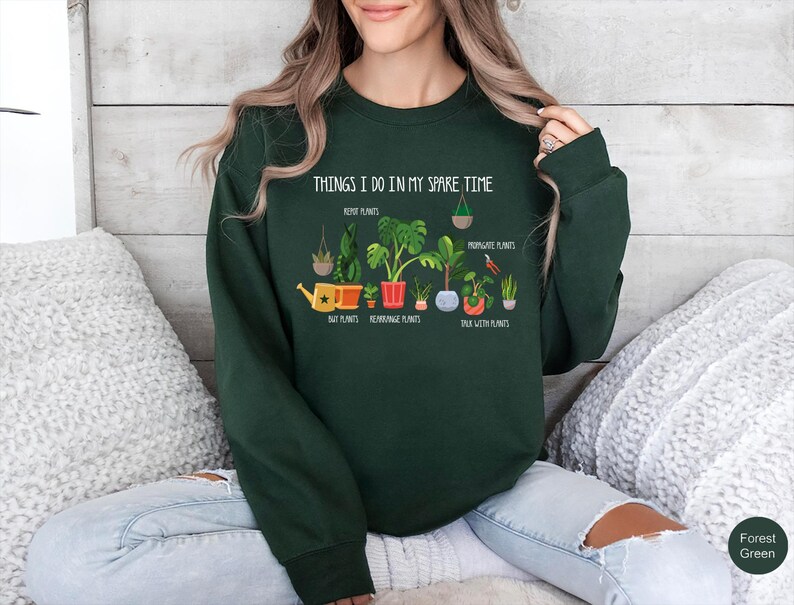 May include: Forest green sweatshirt with the text "THINGS I DO IN MY SPARE TIME" and illustrations of plants. The graphic includes text such as "Repot Plants", "Buy Plants", "Repot Plants", "Propagate Plants", and "Talk With Plants".