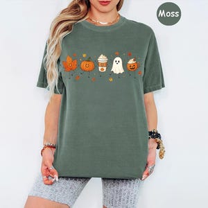 K&ouml;nnte beinhalten: Ein moosgr&uuml;nes Kurzarm-T-Shirt mit einem Grafikdruck, der ein l&auml;chelndes Herbstblatt, einen K&uuml;rbis, einen Kaffeebecher, ein Gespenst und eine Halloween-Laterne zeigt, alle mit Strichm&auml;nnchen-Armen und -Beinen sowie kleinen Herbstbl&auml;ttern.