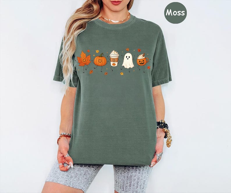 K&ouml;nnte beinhalten: Ein moosgr&uuml;nes Kurzarm-T-Shirt mit einem Grafikdruck, der ein l&auml;chelndes Herbstblatt, einen K&uuml;rbis, einen Kaffeebecher, ein Gespenst und eine Halloween-Laterne zeigt, alle mit Strichm&auml;nnchen-Armen und -Beinen sowie kleinen Herbstbl&auml;ttern.