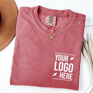 May include: A dusty rose crew neck t-shirt with a Comfort Colors brand label at the inner neckline. The shirt features a white graphic on the lower right side with the text "YOUR LOGO HERE" flanked by two arrows and two horizontal lines.