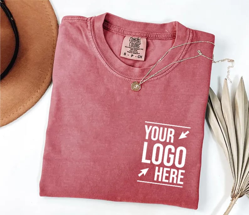 May include: A dusty rose crew neck t-shirt with a Comfort Colors brand label at the inner neckline. The shirt features a white graphic on the lower right side with the text "YOUR LOGO HERE" flanked by two arrows and two horizontal lines.