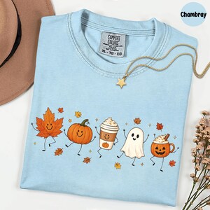 K&ouml;nnte beinhalten: Ein hellblaues T-Shirt mit Rundhalsausschnitt und einem Grafikdruck von tanzendem Ahornblatt, K&uuml;rbis, Kaffeebecher, Geist und Jack-o'-Lantern. Das Shirt hat ein Comfort Colors-Etikett am Halsausschnitt und wird mit einer goldenen Sternkette kombiniert.