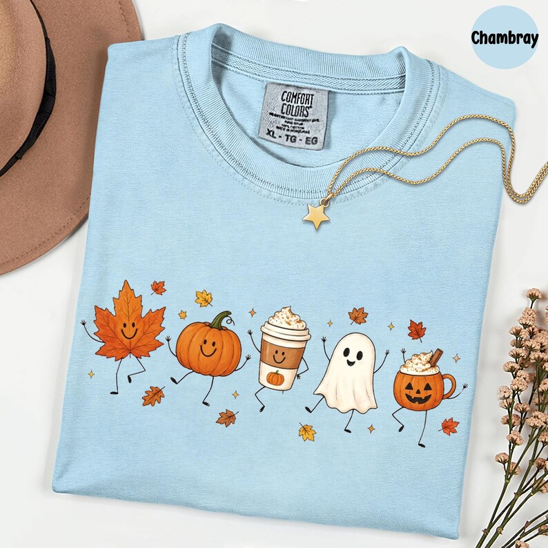 K&ouml;nnte beinhalten: Ein hellblaues T-Shirt mit Rundhalsausschnitt und einem Grafikdruck von tanzendem Ahornblatt, K&uuml;rbis, Kaffeebecher, Geist und Jack-o'-Lantern. Das Shirt hat ein Comfort Colors-Etikett am Halsausschnitt und wird mit einer goldenen Sternkette kombiniert.
