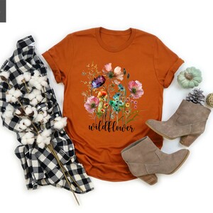 May include: An orange t-shirt with a floral design and the word "wildflower" in black script. The shirt is paired with a black and white plaid shirt, gray boots, and decorative items.