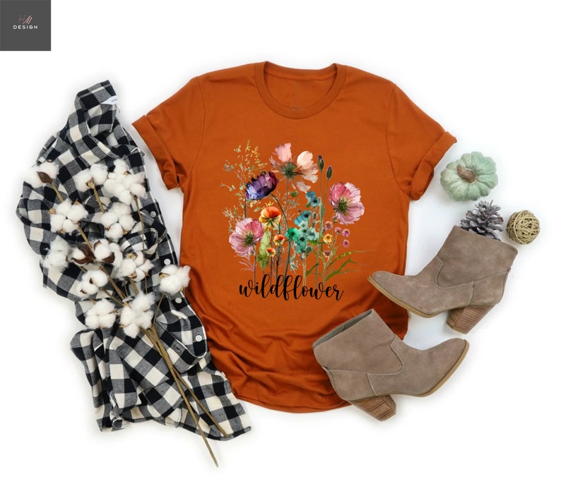 May include: An orange t-shirt with a floral design and the word "wildflower" in black script. The shirt is paired with a black and white plaid shirt, gray boots, and decorative items.