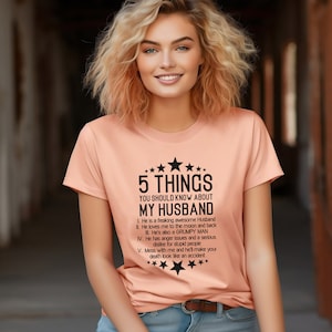 May include: Peach-colored t-shirt with the text "5 THINGS YOU SHOULD KNOW ABOUT MY HUSBAND." The text is in black and includes a list of characteristics. The shirt has a crew neck and short sleeves.