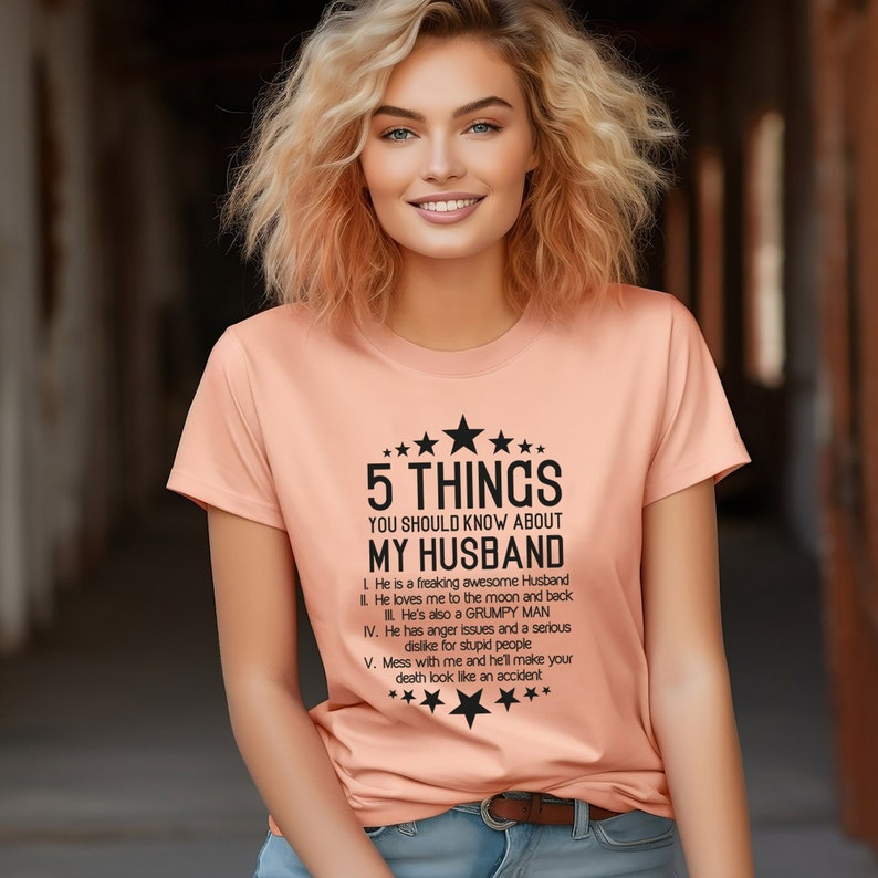 May include: Peach-colored t-shirt with the text "5 THINGS YOU SHOULD KNOW ABOUT MY HUSBAND." The text is in black and includes a list of characteristics. The shirt has a crew neck and short sleeves.