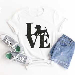 May include: White t-shirt with a black "LOVE" graphic featuring a dachshund silhouette. The shirt is styled with gray sneakers and denim shorts. The design has a distressed, vintage look.