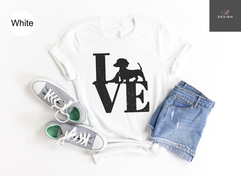 May include: White t-shirt with a black "LOVE" graphic featuring a dachshund silhouette. The shirt is styled with gray sneakers and denim shorts. The design has a distressed, vintage look.
