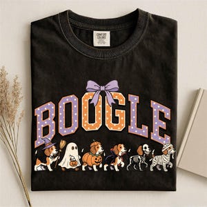 K&ouml;nnte beinhalten: Ein schwarzes Kurzarm-T-Shirt mit dem Schriftzug BOOGLE in gro&szlig;en orangefarbenen und violetten gepunkteten Buchstaben, gekr&ouml;nt von einer violetten Schleife. Darunter befinden sich sechs Beagle-Hunde in Halloween-Kost&uuml;men: Hexe, Geist, K&uuml;rbis, Vampir, Skelett und Mumie.