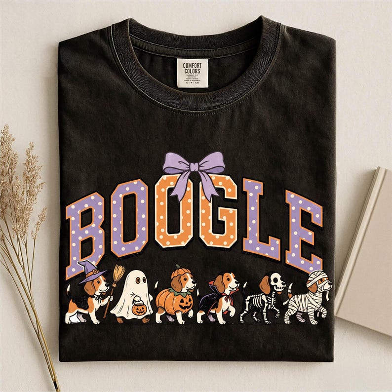K&ouml;nnte beinhalten: Ein schwarzes Kurzarm-T-Shirt mit dem Schriftzug BOOGLE in gro&szlig;en orangefarbenen und violetten gepunkteten Buchstaben, gekr&ouml;nt von einer violetten Schleife. Darunter befinden sich sechs Beagle-Hunde in Halloween-Kost&uuml;men: Hexe, Geist, K&uuml;rbis, Vampir, Skelett und Mumie.