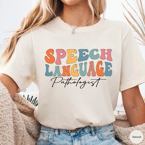 May include: Cream-colored t-shirt with the words "SPEECH LANGUAGE Pathologist" in colorful, retro-style lettering. The text is in orange, pink, yellow, teal, and blue. The shirt is a casual, everyday wear item.