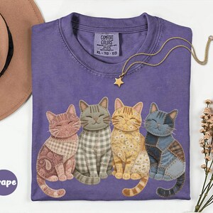 May include: Purple Comfort Colors t-shirt featuring a graphic of four cats in different patterns. A gold necklace with a star pendant is on the shirt. The shirt is size XL-TG-EG.