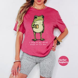 May include: A heather raspberry short-sleeve t-shirt featuring a graphic of a green frog holding a yellow mug. Below the frog, the text reads: i have to say weird stuff or i'll die. The shirt has a crew neckline and a relaxed fit design.