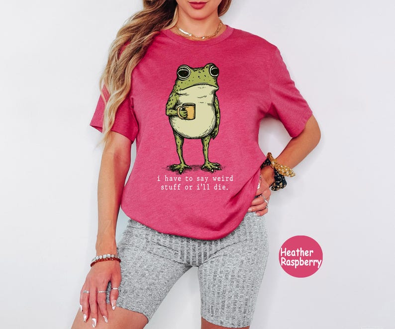 May include: A heather raspberry short-sleeve t-shirt featuring a graphic of a green frog holding a yellow mug. Below the frog, the text reads: i have to say weird stuff or i'll die. The shirt has a crew neckline and a relaxed fit design.