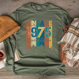 May include: Heather military green long-sleeve shirt with a vintage 1975 graphic in orange, blue, and yellow. A mustard-colored beanie and a plaid scarf are next to the shirt. The shirt is made of a soft, comfortable material.
