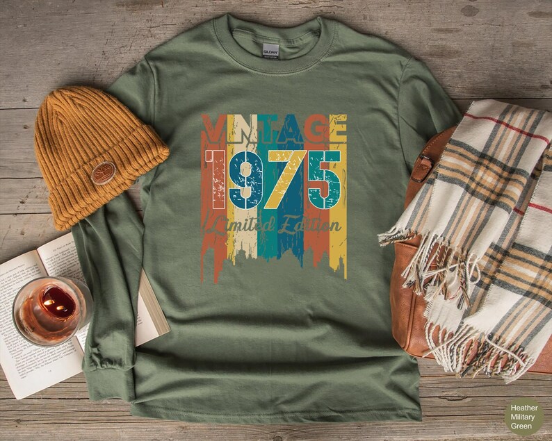 May include: Heather military green long-sleeve shirt with a vintage 1975 graphic in orange, blue, and yellow. A mustard-colored beanie and a plaid scarf are next to the shirt. The shirt is made of a soft, comfortable material.