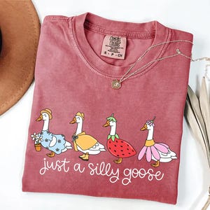 May include: A maroon Comfort Colors t-shirt featuring a whimsical design of four geese dressed in various outfits, including a floral dress, a yellow cape, a strawberry costume, and a pink flower dress. The text "just a silly goose" is printed below the geese.