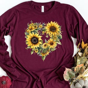 Pode incluir: Camiseta de manga comprida bord&ocirc; com um design floral em forma de cora&ccedil;&atilde;o composto por girass&oacute;is amarelos e folhas verdes. A camiseta tem gola redonda. A palavra "Maroon" est&aacute; impressa no canto inferior direito.