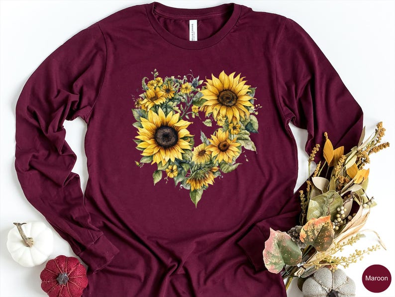 Pode incluir: Camiseta de manga comprida bord&ocirc; com um design floral em forma de cora&ccedil;&atilde;o composto por girass&oacute;is amarelos e folhas verdes. A camiseta tem gola redonda. A palavra "Maroon" est&aacute; impressa no canto inferior direito.