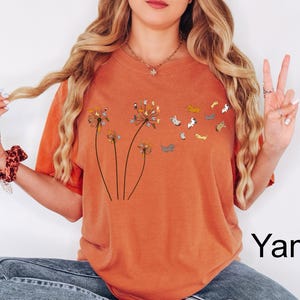 May include: A burnt orange t-shirt featuring a graphic of three dandelion stalks where the seeds are replaced by small, colourful illustrated cats. The cats appear to be floating away from the dandelion heads in a whimsical, scattered pattern.
