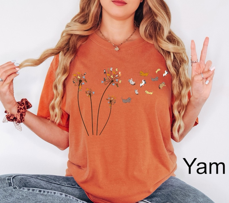 May include: A burnt orange t-shirt featuring a graphic of three dandelion stalks where the seeds are replaced by small, colourful illustrated cats. The cats appear to be floating away from the dandelion heads in a whimsical, scattered pattern.