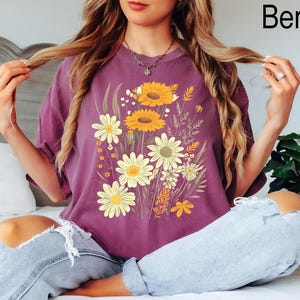 K&ouml;nnte beinhalten: Beerenfarbenes T-Shirt mit einem Blumendesign mit Sonnenblumen und G&auml;nsebl&uuml;mchen. Die Grafik enth&auml;lt gelbe, wei&szlig;e und gr&uuml;ne Blumen. Das Shirt hat eine lockere Passform.