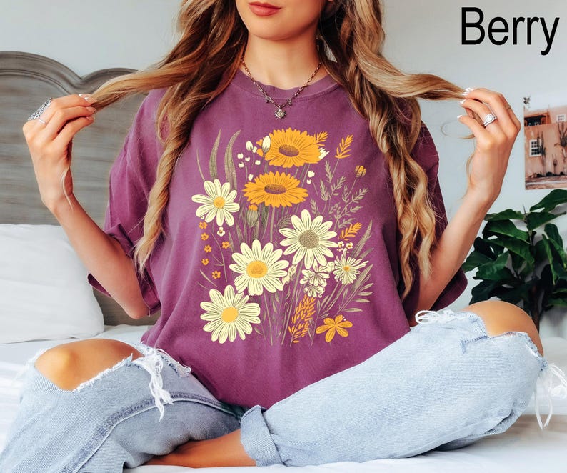 K&ouml;nnte beinhalten: Beerenfarbenes T-Shirt mit einem Blumendesign mit Sonnenblumen und G&auml;nsebl&uuml;mchen. Die Grafik enth&auml;lt gelbe, wei&szlig;e und gr&uuml;ne Blumen. Das Shirt hat eine lockere Passform.