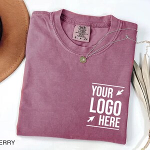 May include: A berry-colored short-sleeve t-shirt with a crew neckline and a Comfort Colors brand label at the inner collar. The front features white text that reads "YOUR LOGO HERE" framed by two horizontal lines and two small arrows.