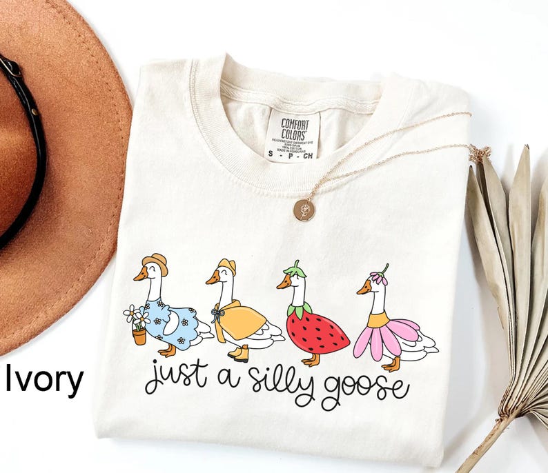 May include: Ivory-colored t-shirt featuring a whimsical design of four geese dressed in various outfits, including a dress, raincoat, strawberry costume, and flower dress. The phrase "just a silly goose" is written in a playful script below the geese.