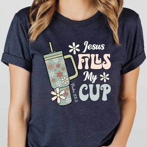 K&ouml;nnte beinhalten: Dunkelblaues T-Shirt mit dem Text "Jesus Fills My Cup" in Retro-Schrift, zusammen mit einem floral gemusterten Becher. Das Design enth&auml;lt auch den Bibelvers Psalm 23:5. Das T-Shirt hat eine dunkelblaue Farbe.