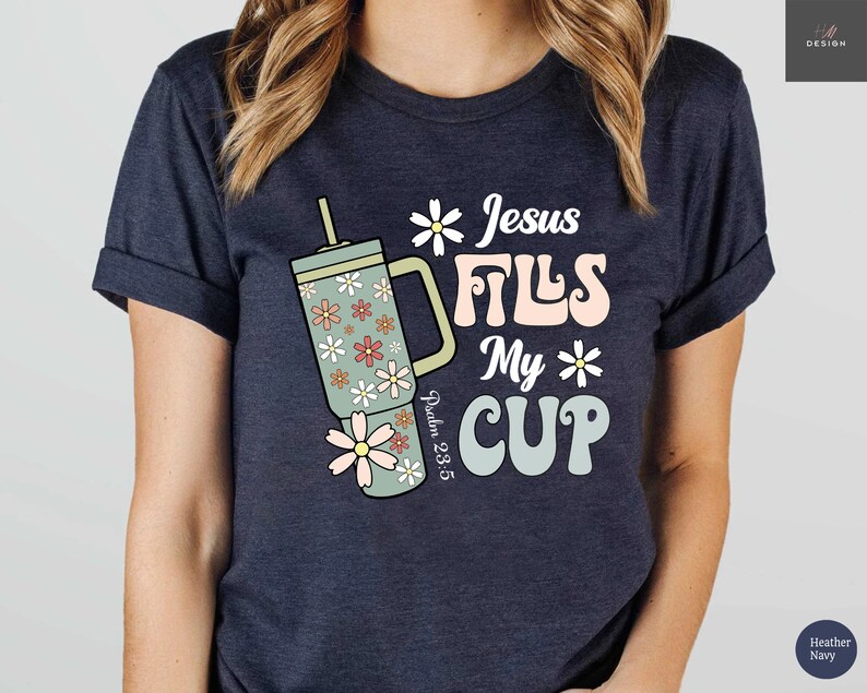 K&ouml;nnte beinhalten: Dunkelblaues T-Shirt mit dem Text "Jesus Fills My Cup" in Retro-Schrift, zusammen mit einem floral gemusterten Becher. Das Design enth&auml;lt auch den Bibelvers Psalm 23:5. Das T-Shirt hat eine dunkelblaue Farbe.