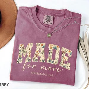 May include: Berry-colored t-shirt with the words "MADE for more" in a floral design. The text "Ephesians 2:10" is below the main text. A silver necklace with a pendant is on the shirt. The shirt is folded and next to a hat and dried plant.