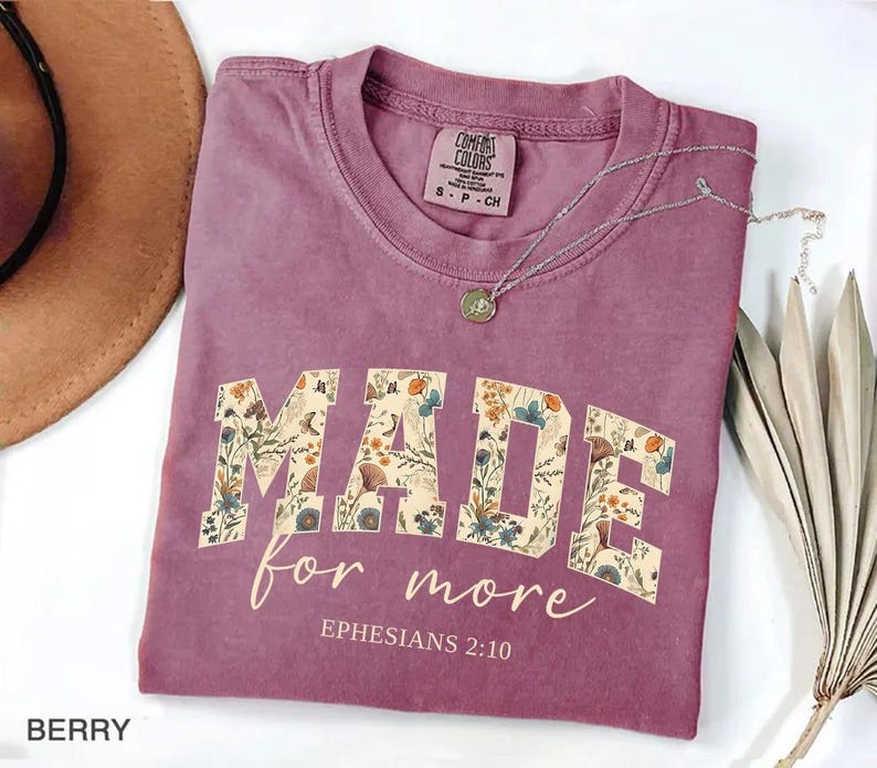 May include: Berry-colored t-shirt with the words "MADE for more" in a floral design. The text "Ephesians 2:10" is below the main text. A silver necklace with a pendant is on the shirt. The shirt is folded and next to a hat and dried plant.