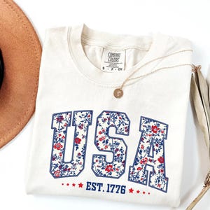 May include: Cream-colored t-shirt with the word "USA" in a floral pattern, outlined in blue. The text "EST. 1776" is below, with red stars. A gold necklace and a brown hat are also visible.
