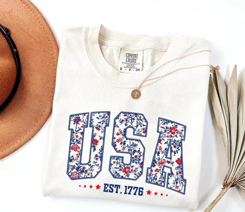 May include: Cream-colored t-shirt with the word "USA" in a floral pattern, outlined in blue. The text "EST. 1776" is below, with red stars. A gold necklace and a brown hat are also visible.