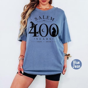 Puede incluir: Una camiseta de manga corta color azul jean con un estampado gr&aacute;fico negro que dice SALEM MASSACHUSETTS 400 YEARS 1626-2026. El dise&ntilde;o incluye un sombrero de bruja, una luna creciente y la silueta de un gato integrados en el n&uacute;mero 400.
