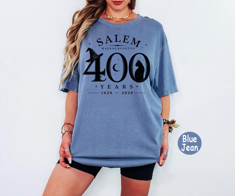 Puede incluir: Una camiseta de manga corta color azul jean con un estampado gr&aacute;fico negro que dice SALEM MASSACHUSETTS 400 YEARS 1626-2026. El dise&ntilde;o incluye un sombrero de bruja, una luna creciente y la silueta de un gato integrados en el n&uacute;mero 400.