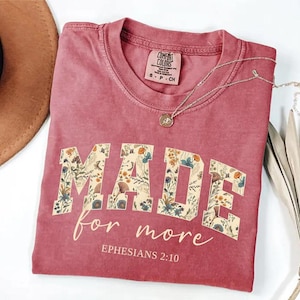 May include: A dusty rose Comfort Colors t-shirt with the words "MADE for more" in a floral design. The text "Ephesians 2:10" is below the main text. A silver necklace with a pendant is on the shirt.