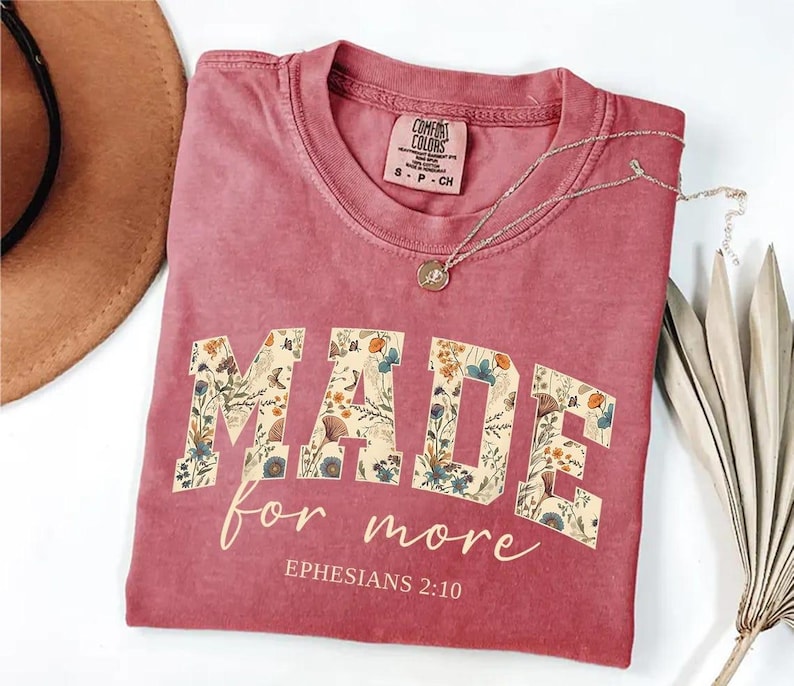 May include: A dusty rose Comfort Colors t-shirt with the words "MADE for more" in a floral design. The text "Ephesians 2:10" is below the main text. A silver necklace with a pendant is on the shirt.