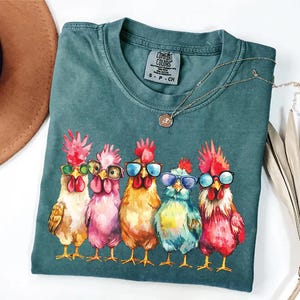 Puede incluir: Una camiseta azul verdosa con un gr&aacute;fico de cinco pollos coloridos que llevan gafas de sol. Los pollos est&aacute;n dispuestos en fila, con la cabeza girada hacia un lado. La camiseta tiene el texto "Comfort Colors" impreso en la etiqueta.