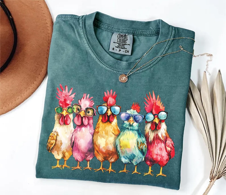 Puede incluir: Una camiseta azul verdosa con un gr&aacute;fico de cinco pollos coloridos que llevan gafas de sol. Los pollos est&aacute;n dispuestos en fila, con la cabeza girada hacia un lado. La camiseta tiene el texto "Comfort Colors" impreso en la etiqueta.