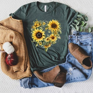 Pode incluir: T-shirt verde escuro com um design de girass&oacute;is em forma de cora&ccedil;&atilde;o. A imagem inclui jeans azuis, botins castanhos e um cardigan bege. Uma ab&oacute;bora decorativa e uma bola de l&atilde; tamb&eacute;m est&atilde;o presentes.