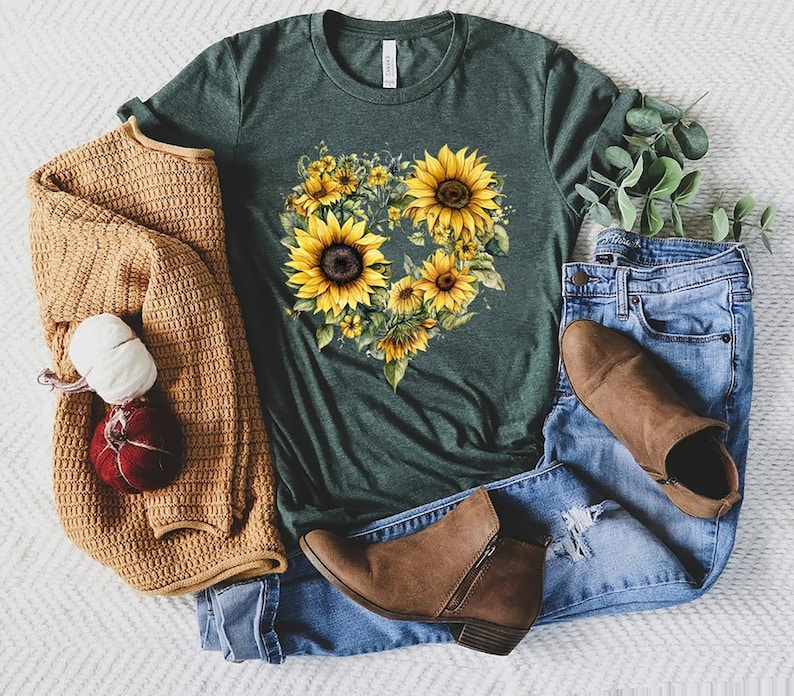 Pode incluir: T-shirt verde escuro com um design de girass&oacute;is em forma de cora&ccedil;&atilde;o. A imagem inclui jeans azuis, botins castanhos e um cardigan bege. Uma ab&oacute;bora decorativa e uma bola de l&atilde; tamb&eacute;m est&atilde;o presentes.