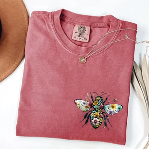 May include: A dusty rose Comfort Colors t-shirt with a floral bee graphic. The bee design features colorful flowers and leaves. A gold necklace with a small pendant rests on the shirt. The shirt has a small Comfort Colors tag.
